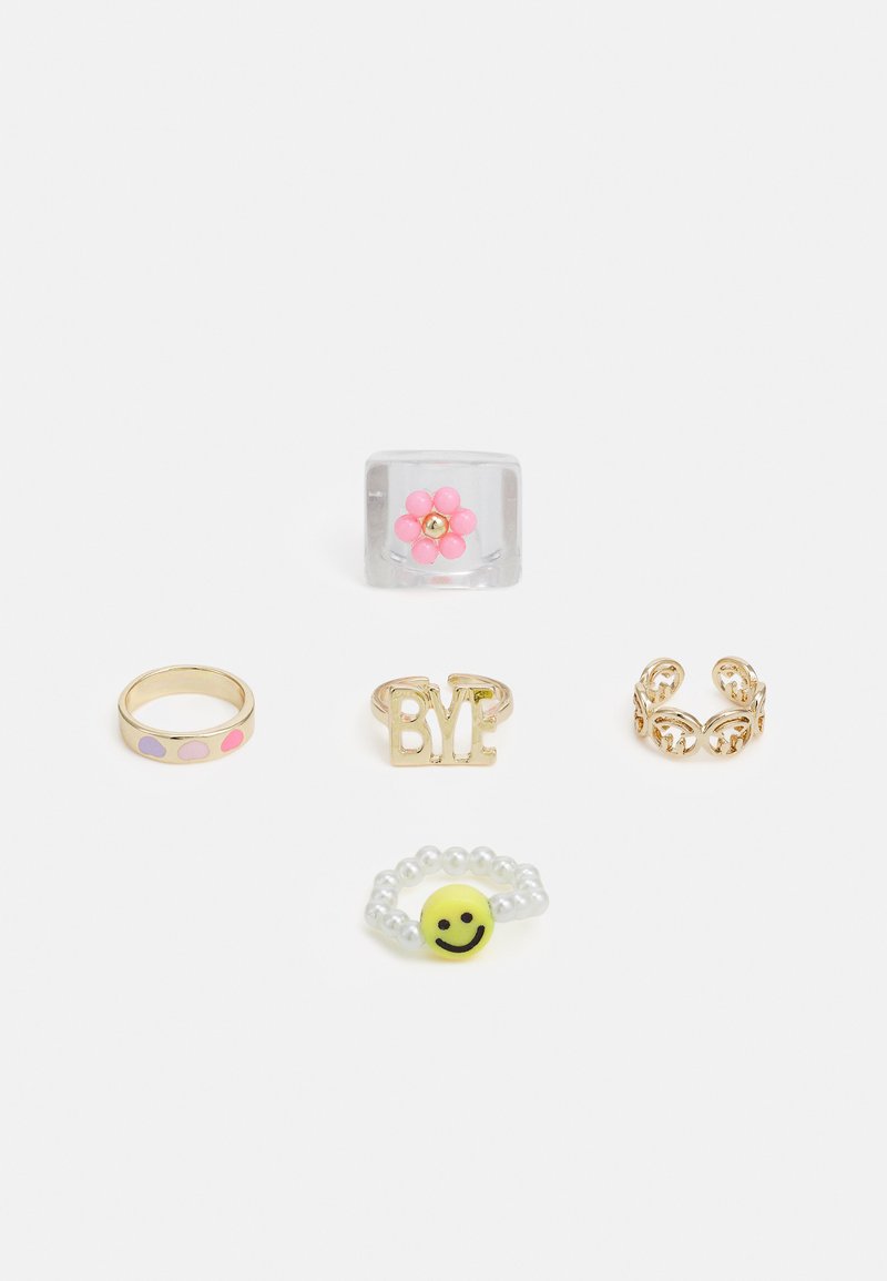 Cinco anéis exibidos: um anel de flor transparente, um anel dourado com a palavra "BYE", um anel dourado com pontos coloridos, um anel de filigrana dourada e um anel de contas com um sorriso.