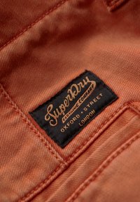 Pomarańczowa tkanina z widocznymi szwami ma czarną metkę z napisem "SuperDry Clothing Company" oraz "Oxford Street, Londyn."