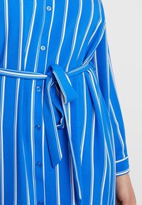 Abito camicia a righe blu e bianche con vita regolabile, dotato di chiusura frontale con bottoni e maniche lunghe, realizzato in tessuto morbido.