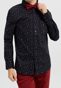 Camisa negra con pequeños patrones geométricos blancos, rojos y azules, de mangas largas y con cierre de botones. Combinada con una pajarita roja.