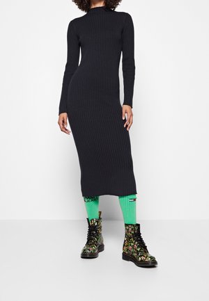 Prążkowana, czarna sukienka z długim rękawem i wysokim kołnierzem, połączona z jasnymi zielonymi skarpetkami i botkami w kwiatowy wzór, prezentująca unikalne tekstury i kolory.