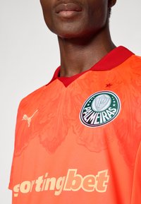 Oranžna športna majica z rdečim ovratnikom, ki vsebuje podroben vzorec leva, zeleno logotip Palmeiras in "sportingbet" v krepkem rumenem besedilu.