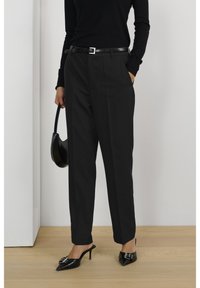 Pantaloni neri su misura con vita alta, tasche laterali e design elegante, abbinati a mule nere con tacco alto e una borsa.