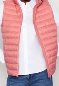 Rosa Steppweste mit Reißverschluss, gestepptem Material und hohem Kragen. Getragen über einem weißen Hemd mit kleinem Logo auf der Brust.