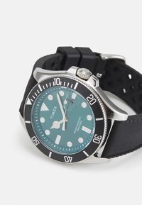Reloj Timex con esfera color teal, bisel negro, caja plateada y correa de goma. Presenta índices de hora y una ventana de fecha a las 3 en punto. Resistente al agua.