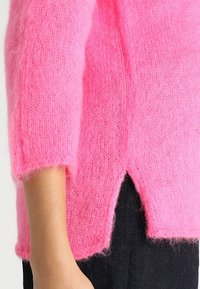 Jasně růžový pletený svetr s krátkými rukávy, disponující měkkou, nadýchanou texturou a bočními rozparky na lemu pro snadnější pohyb.