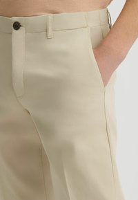 Pantalones beiges de corte recto, confeccionados en una tela suave. Cuentan con cierre de botones y bolsillos laterales, con detalles de costura visibles.