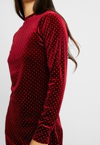 Röd sammetklänning med långa ärmar, med en åtsittande siluett och spridda silverprickar över tyget. Texturerad yta.