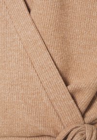 Primo piano di tessuto beige a maglia coste con design incrociato a fascia e dettaglio cintura annodata.