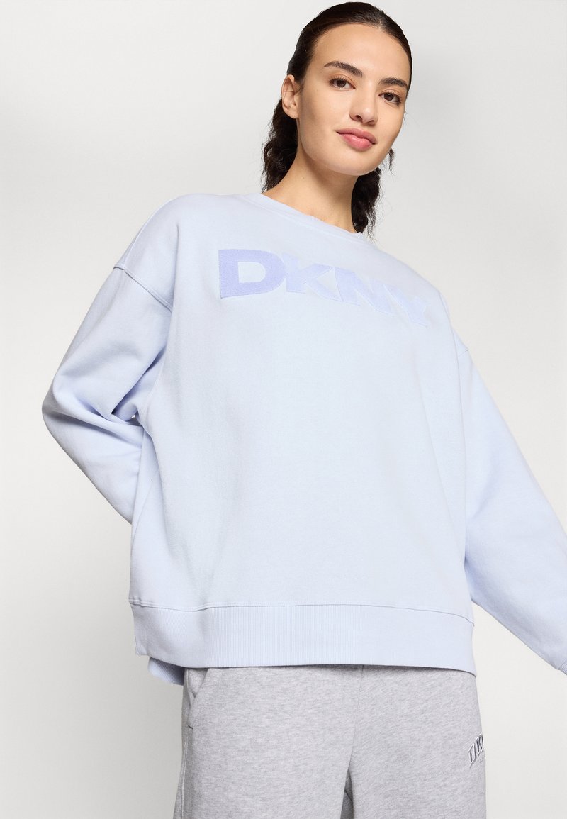 Jasnoniebieska sweatshirt z logo DKNY o miękkiej fakturze. Posiada obniżone ramiona, ściągacze oraz luźny krój. Zestawiona z szarymi dresami.