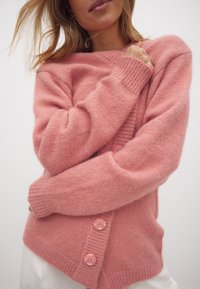 Różowy sweter z dzianiny wykonany z miękkiego, puszystego materiału, z okrągłym dekoltem, ściągaczami przy mankietach i trzema różowymi guzikami z przodu.