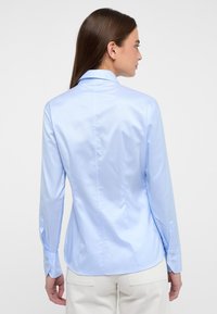 Camicia azzurra chiara a maniche lunghe realizzata in tessuto morbido, con colletto classico e polsini con bottoni, design aderente con dettagli cuciti sul retro.