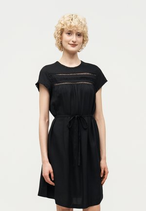 Kobieta o krótkich kręconych blond włosach, ubrana w czarną sukienkę do kolan z koronkowymi detalami i wiązaniem w talii, stojąca na jednolitym białym tle.