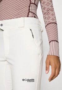 Biele nohavice Columbia Titanium so slim fit strihom, ktoré majú zipsovú kapsu a detail loga na stehne; materiál vyzerá pružný a hladký.