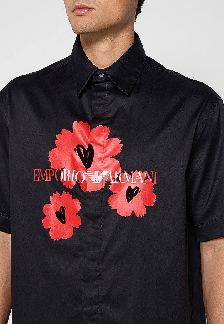 Camisa negra de manga corta con tres grandes flores rojas y detalles de corazones negros, con el logo de Emporio Armani en blanco y rojo en el pecho.
