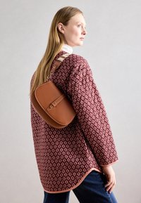 Bruine leren schoudertas met een afgeronde vorm, voorzien van een sluiting met gesp en een gestructureerde band, gedragen over een bordeauxrood shirt met patroon.