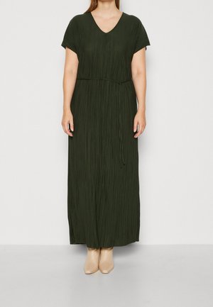 Femme portant une robe maxi plissée vert foncé à manches courtes avec un col en V et des bottines beige, debout devant un fond uni.