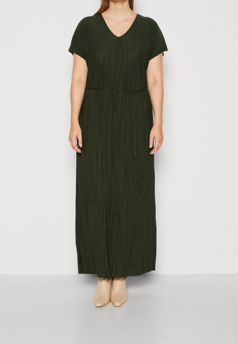 Femme portant une robe maxi plissée vert foncé à manches courtes avec un col en V et des bottines beige, debout devant un fond uni.