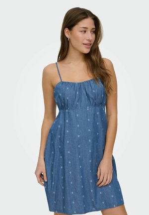 Tânără purtând o rochie albastră din denim, fără mâneci, cu bretele subțiri și un model paisley subtil, uitându-se spre dreapta.