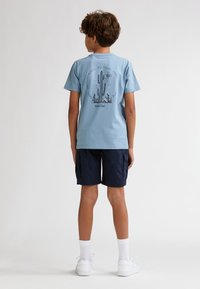 T-shirt en coton bleu clair avec un motif de cactus dans le dos, associé à un short cargo marine. Le mannequin porte des baskets blanches et des chaussettes montantes.