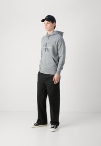 Felpa grigia con cappuccio e coulisse, logo frontale; abbinata a pantaloni neri. Modello che indossa un cappellino blu e scarpe nere con dettagli bianchi.