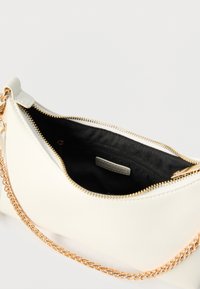 Petit sac à main crème avec une bandoulière chaîne dorée, ouverture du dessus zippée laissant apparaître un intérieur en tissu noir et une poche intérieure zippée.