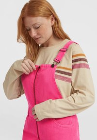 Rødhåret kvinne i beige genser med striper som åpner fronten av lys rosa kordfløyelsdresser mot hvit bakgrunn.