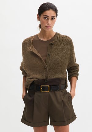 Femme aux cheveux foncés attachés en arrière, portant un cardigan marron texturé sur un chemisier marron foncé et un short marron taille haute avec une ceinture noire.