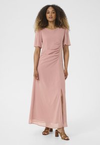 Long pink dress with short sleeves, gathered side detail, and a thigh-high slit. Smooth, flowy fabric. Worn with brown block-heeled sandals.