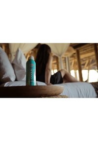 Turkusowa butelka z filtrami przeciwsłonecznymi SPF 50, z białą nakrętką i tropikalnym wzorem, umieszczona w drewnianej misce obok łóżka z białymi prześcieradłami.