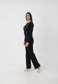 Blusa negra de botones con ribete blanco en forma de ondas, combinada con pantalones anchos negros y tacones abiertos blancos. Diseño minimalista, silueta elegante.