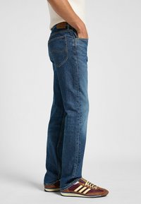 Vaqueros de denim azul con un diseño de pierna recta, cinco bolsillos, costuras visibles y un parche de cuero en la cintura. El calzado presenta acentos en marrón y amarillo.