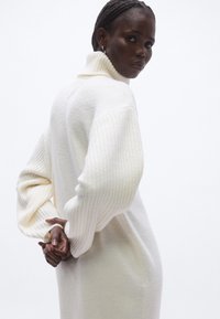 Nagyméretű, bordázott pulóverruha törtfehér színben, magas gallérral és széles ujjakkal. Puha tapintású, laza szabású.