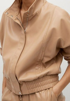 Persona con chaqueta de polipiel color beige con cuello alto y pantalones a juego, mano en el bolsillo, mostrada desde el cuello hasta las caderas.
