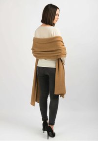 Beige uldtrøje lagdelt med et brunt sjal, båret over mørke skinny jeans og sorte højhælede sko. Ensfarvet baggrund.