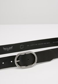 Ceinture en cuir noir avec une boucle ovale en argent, dotée de perforations pour un ajustement et de détails gaufrés à l'intérieur.