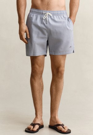 Man draagt blauw-wit geruite zwemshorts met trekkoord en donkerbruine slippers, staand tegen een effen achtergrond.