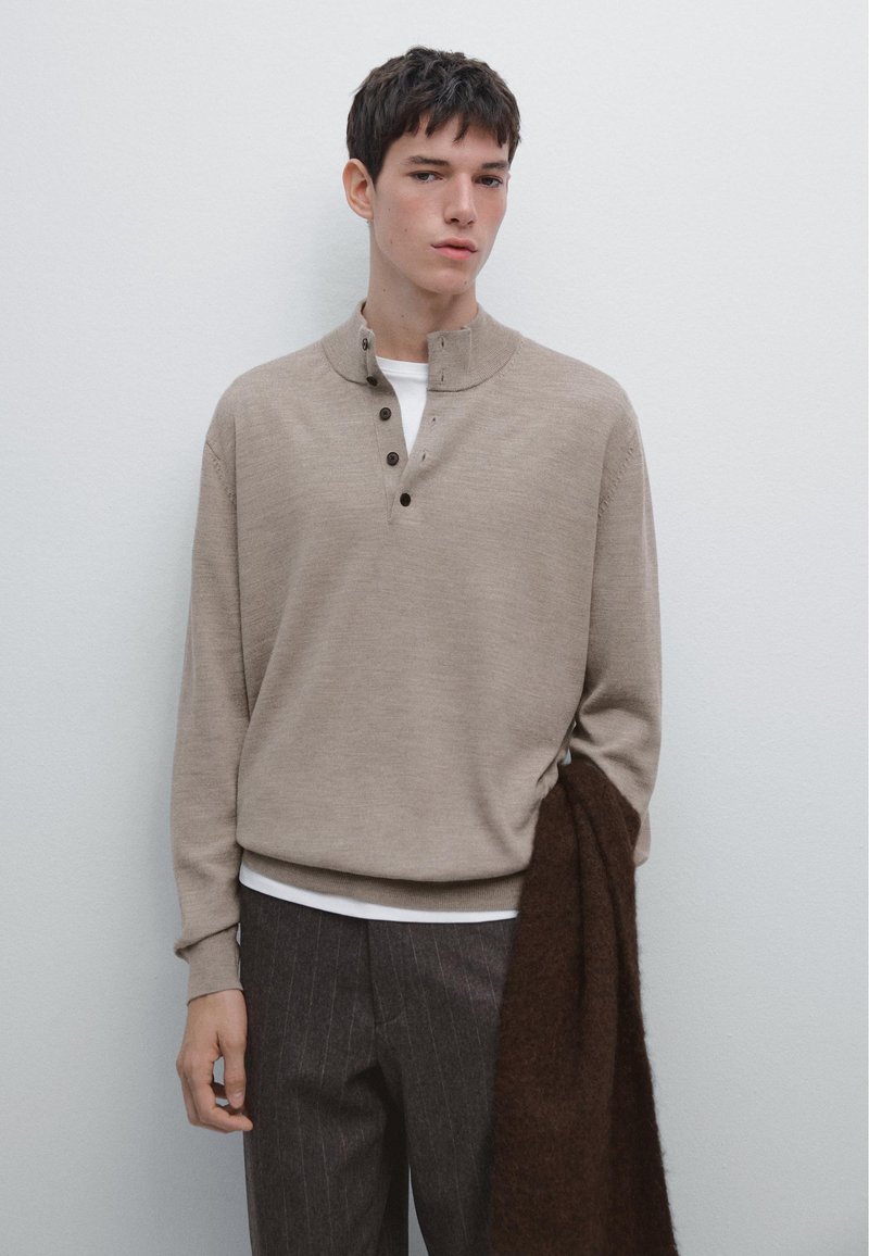 Pullover beige a maglia con colletto alto e tre bottoni marroni. Abbinato a pantaloni a righe sottili e una sciarpa marrone appoggiata sul braccio.
