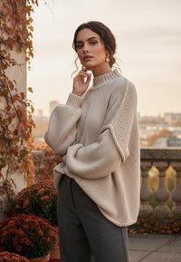 Jasno beżowy sweter dzianinowy z wysokim ściągaczem, szerokimi rękawami i luźnym krojem, zestawiony z ciemnoszarymi spodniami na tle jesiennych liści.