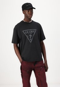 Sort kortærmet T-shirt med et trekantet gråt "GUESS" logo. Kombineret med røde strukturerede bukser og suppleret med en sort kasket.