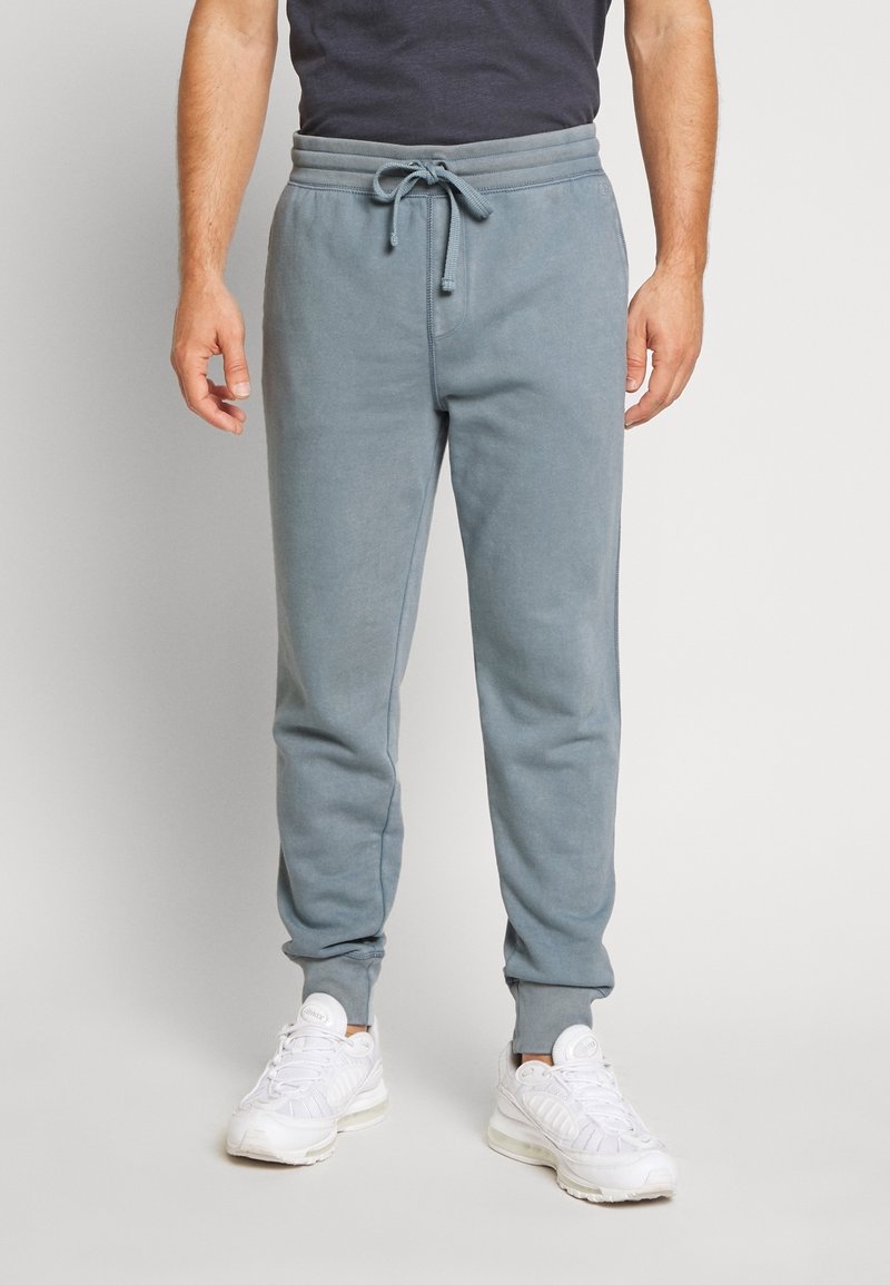 Homme portant un pantalon de jogging bleu clair avec cordon à la taille et des baskets blanches, debout devant un fond neutre.