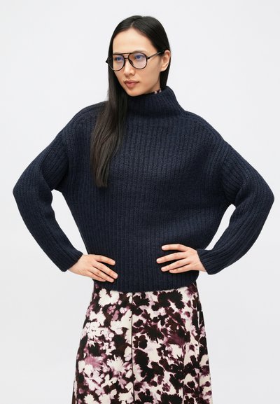 Σκούτερο μπλε πλεκτό πουλόβερ με ψηλό γιακά, σε συνδυασμό με φούστα με λουλουδάτο μοτίβο σε αποχρώσεις του μωβ και του λευκού. Μακριά μανίκια και άνετη εφαρμογή.