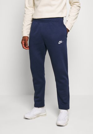 Mężczyzna ubrany w granatowe dresy Nike, białe tenisówki i kremową bluzę z długim rękawem stoi na jednolitym tle.