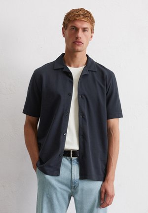 Blond man wearing a dark short-sleeve button-up shirt over a white t-shirt with light blue jeans, standing against a plain white wall.
