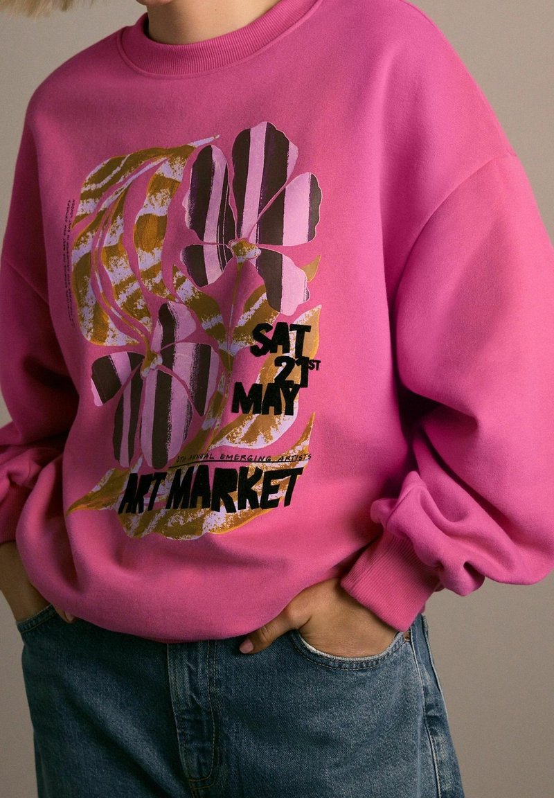 Personne portant un sweat-shirt rose avec un motif floral abstrait et le texte « Sam 21 mai Marché d'Art », les mains dans les poches d'un jean bleu.
