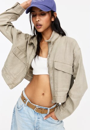 Veste courte kaki avec deux poches avant, tissu texturé et col montant. Associée à un crop top blanc et un jean bleu clair.