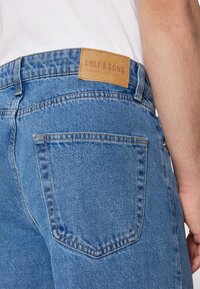 Light blue denim jeans featuring a leather patch on the back waistband with "ONLY & SONS" embossed. Two rear pockets with visible stitching.