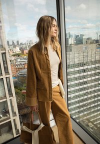 Ženska v rjavem kordurovem kostimu drži usklajeno torbico, stoji ob velikem oknu z razgledom na mestne stavbe na sončen dan.