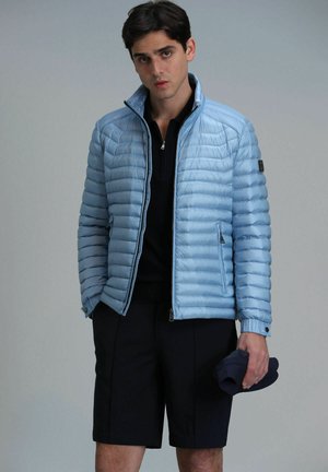 Light blue puffer jacket with horizontal quilted design, zippered pockets, high collar, paired with navy shorts, held cap in hand.