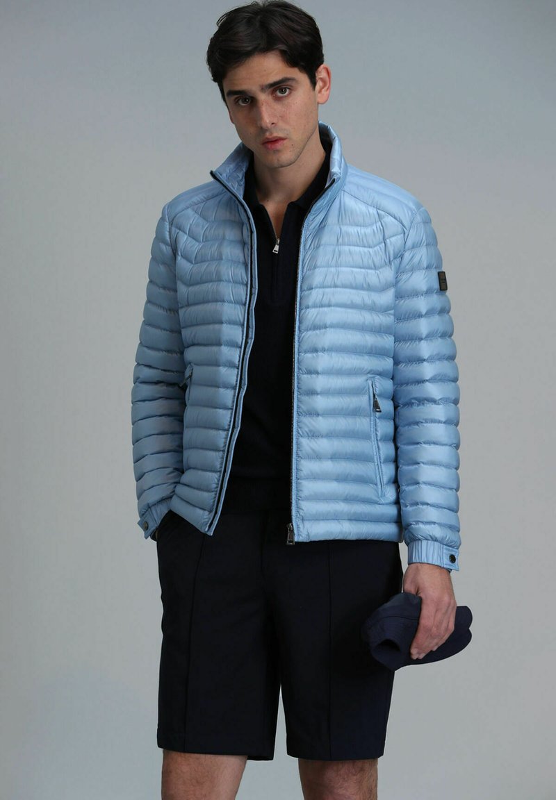 Veste d'hiver légère bleue claire avec un design matelassé horizontal, poches zippées, col montant, associée à un short navy, tenant une casquette à la main.
